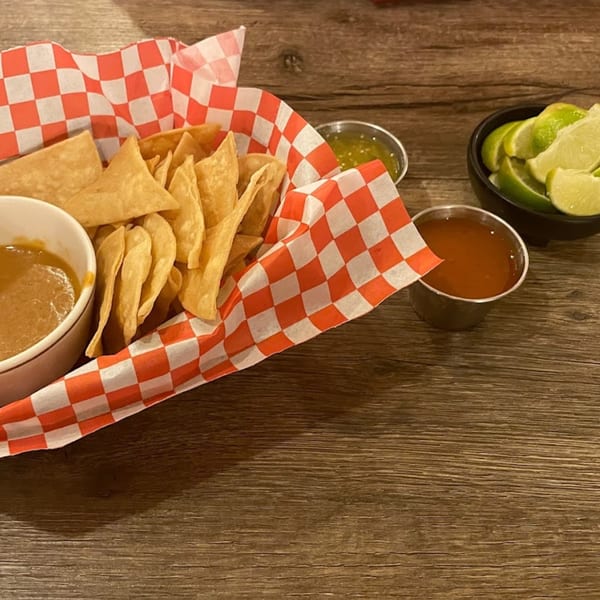 A basket of tortilla chips with consome, salsa verde, red salsa, and lime wedges on a wooden table.