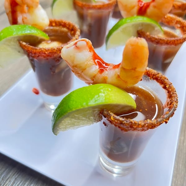 Shrimp cocktail shooters with lime wedges and chili-rimmed glasses on a white tray.