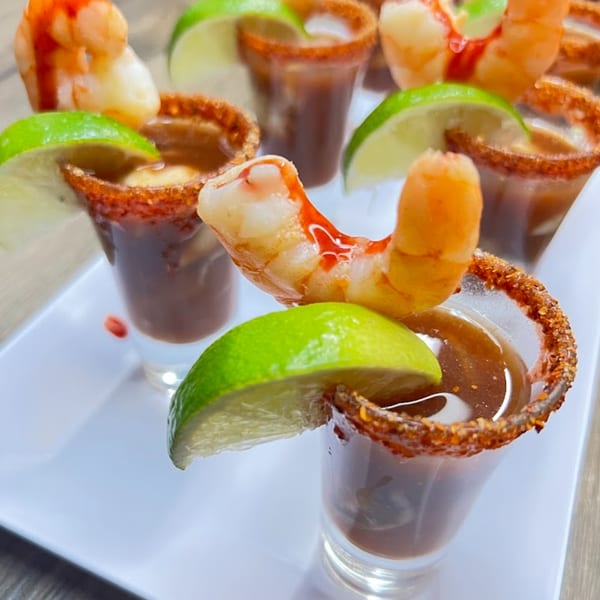 Shrimp cocktail shooters with lime wedges and chili-rimmed glasses on a white tray.