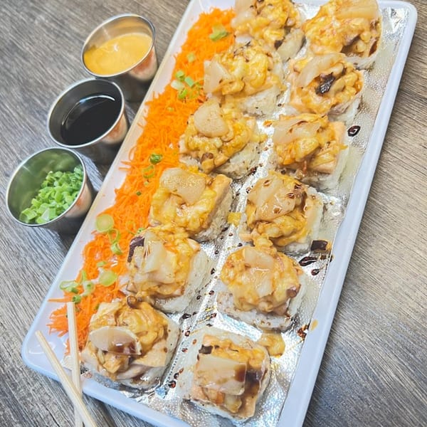 A white platter of baked sushi rolls with sauces and shredded carrots on a wooden table.