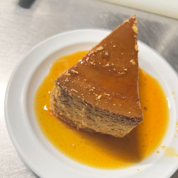 A triangular slice of flan with caramel sauce on a white plate from Birria - Bite los Arcos.