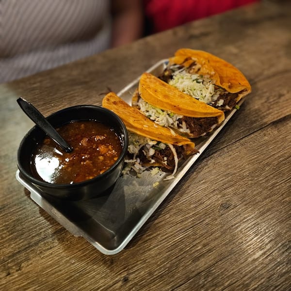 Three Birria tacos with shredded cabbage and a side of consommé on a metal tray.
